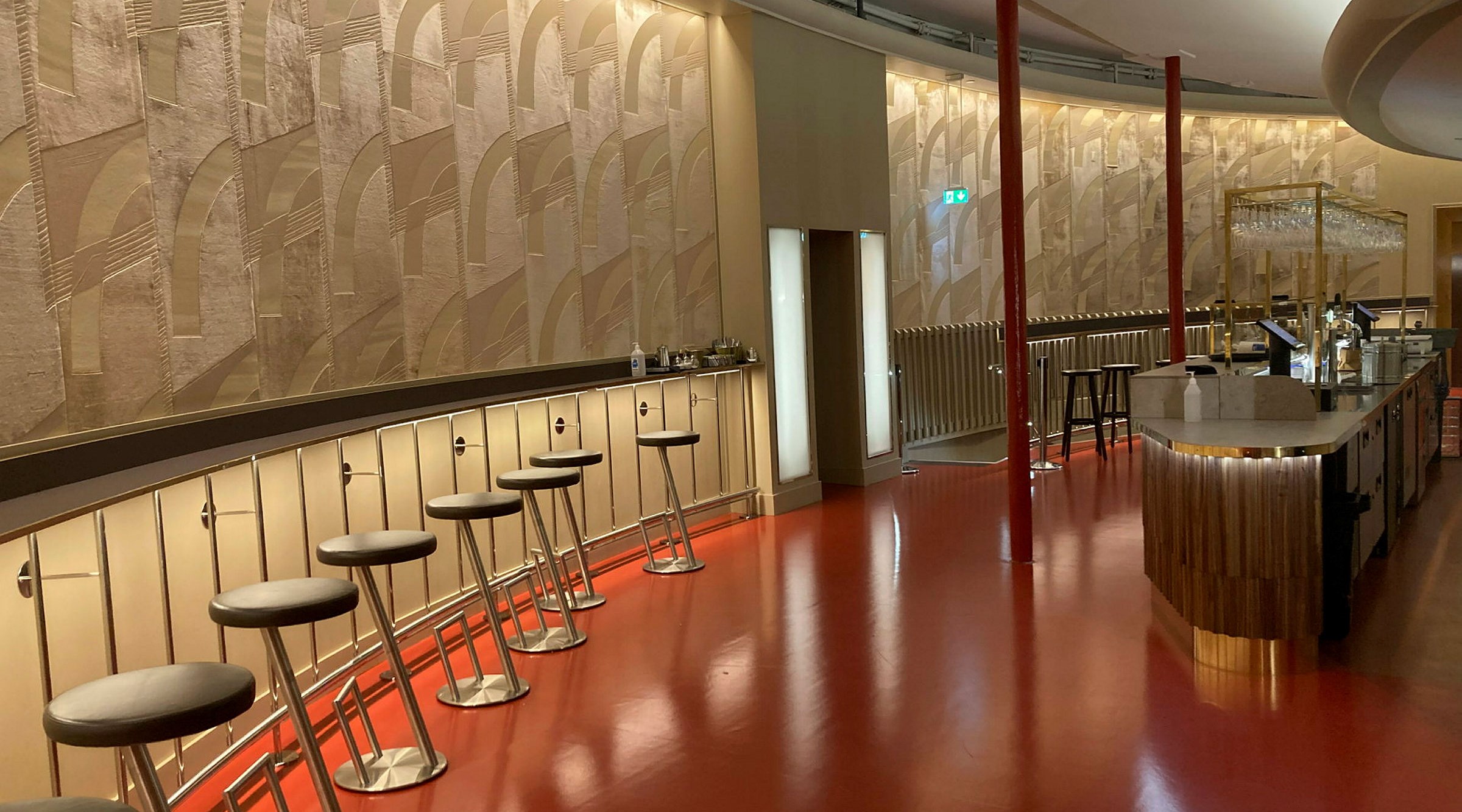 Den nyrenoverade foajébaren på Stadsteatern i Göteborg. en böjd fondvägg klädd med den gyllene tapeten med geometriskt mönster. Utmed väggen löper en väggfast smal borsyta med barstolar placerade med jämna intervall. Till höger i rummet en bardisk klädd i trä. Golvet är rött och högblankt.