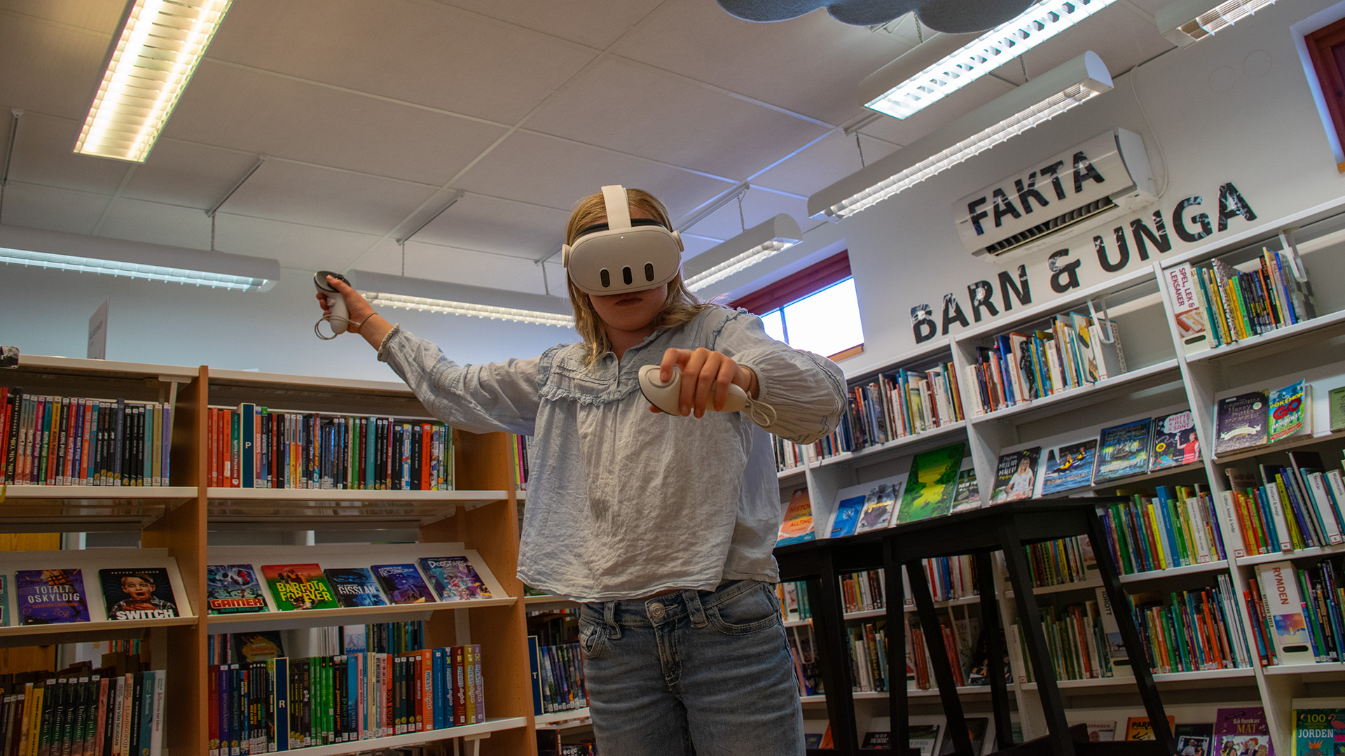 En flicka står med VR-headset inne bland bokhyllorna på barn- och ungdomsavdelningen på biblioteket i Kungshamn. Hon spelar och är i rörelse, i händerna håller hon spelkontroller. Hon har långt blont hår och är klädd i blå jeans och en ljusblå blus. 
