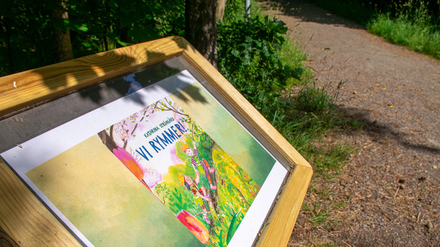En låg skylt med träram vid sidan av en gångväg i skogen. Innanför glaset finns framsidan till boken Vi rymmer.