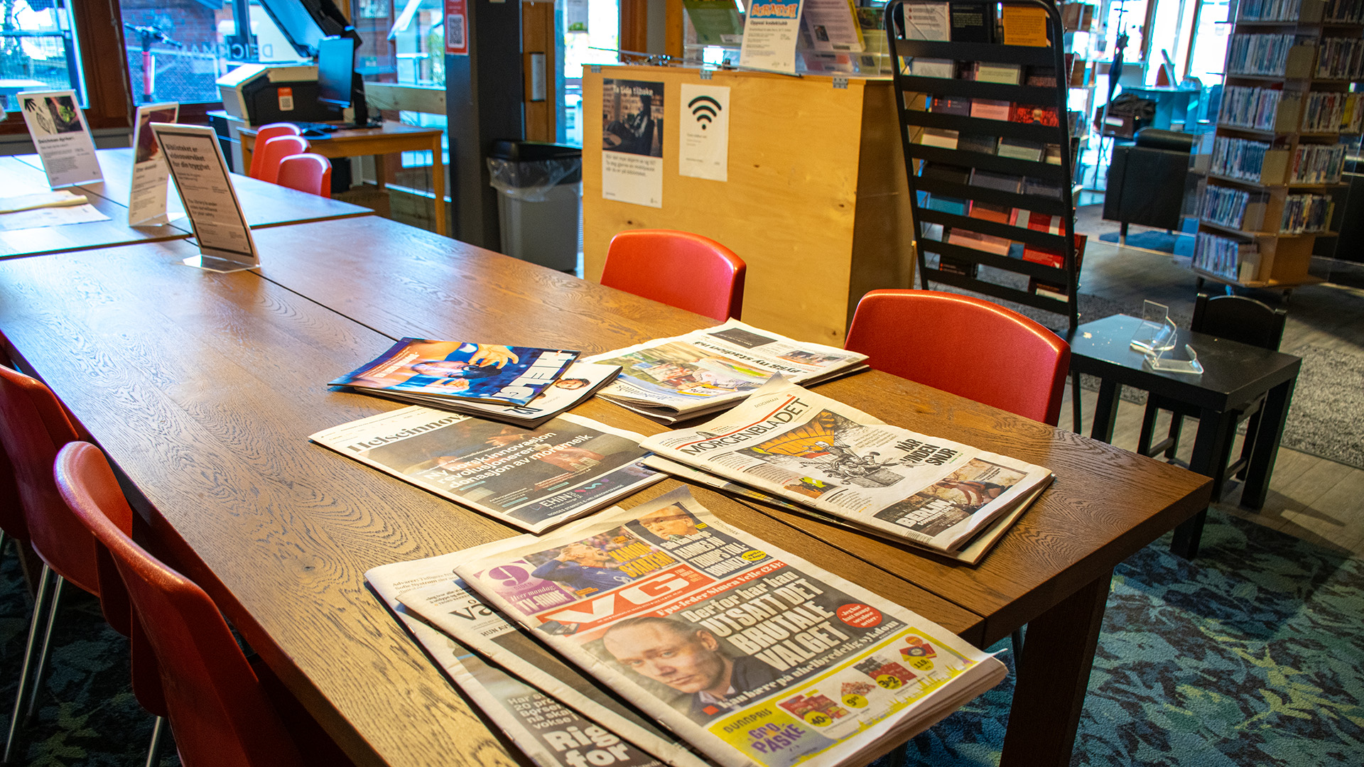 Ett rektangulärt långt bord med röda stolar. På bordet ligger dagstidningar utspridda. Ett litet svart bord med en liten svart stol står till höger. Stora glasrutor från golv till tak släpper in ljus i rummet. 